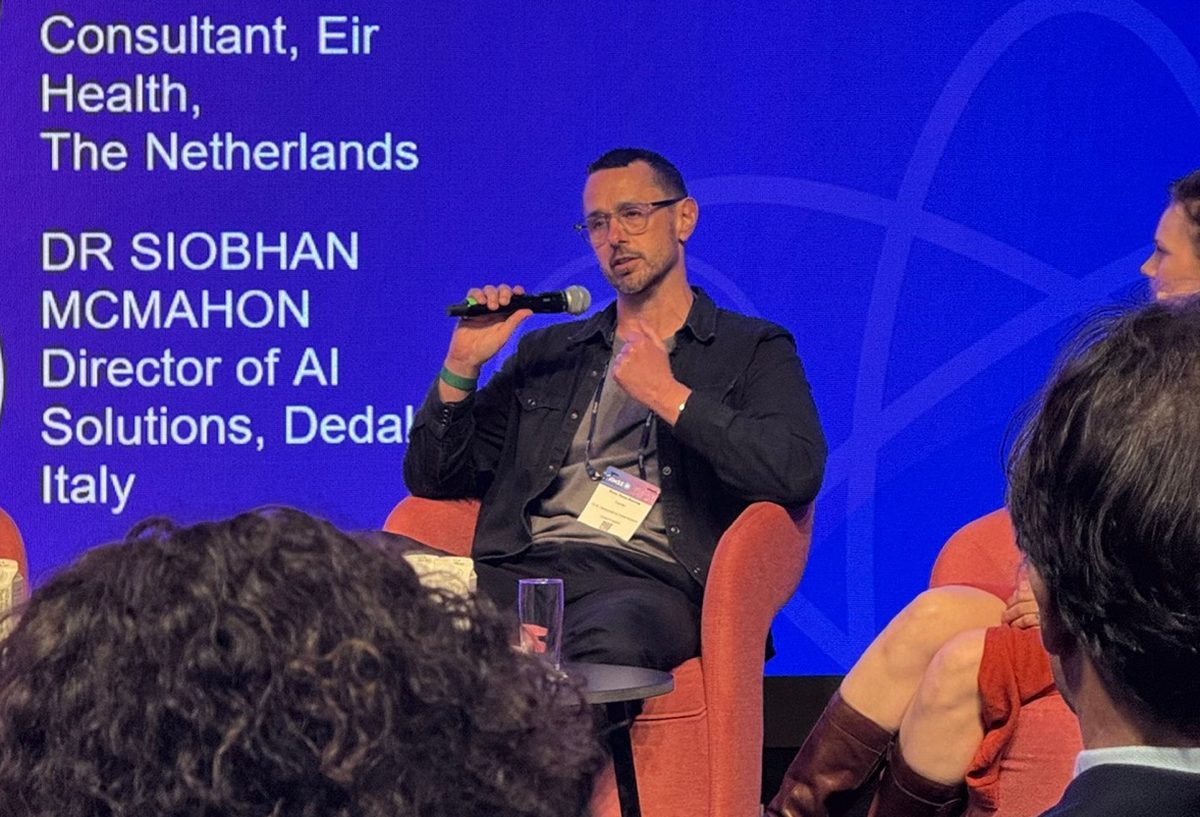 Russ Read-Barrow speaking with a microphone on a conference panel, seated in front of a large blue screen listing the other panellists.